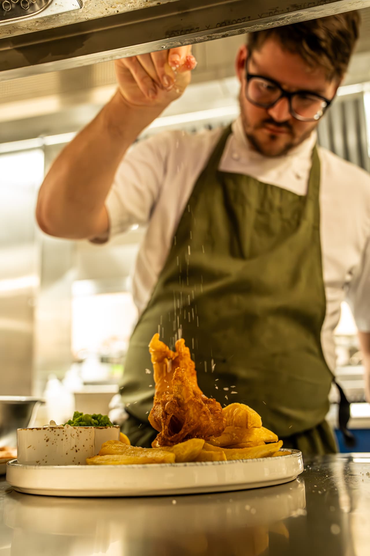 A chef seasoning fish and chips at the kitchen pass
