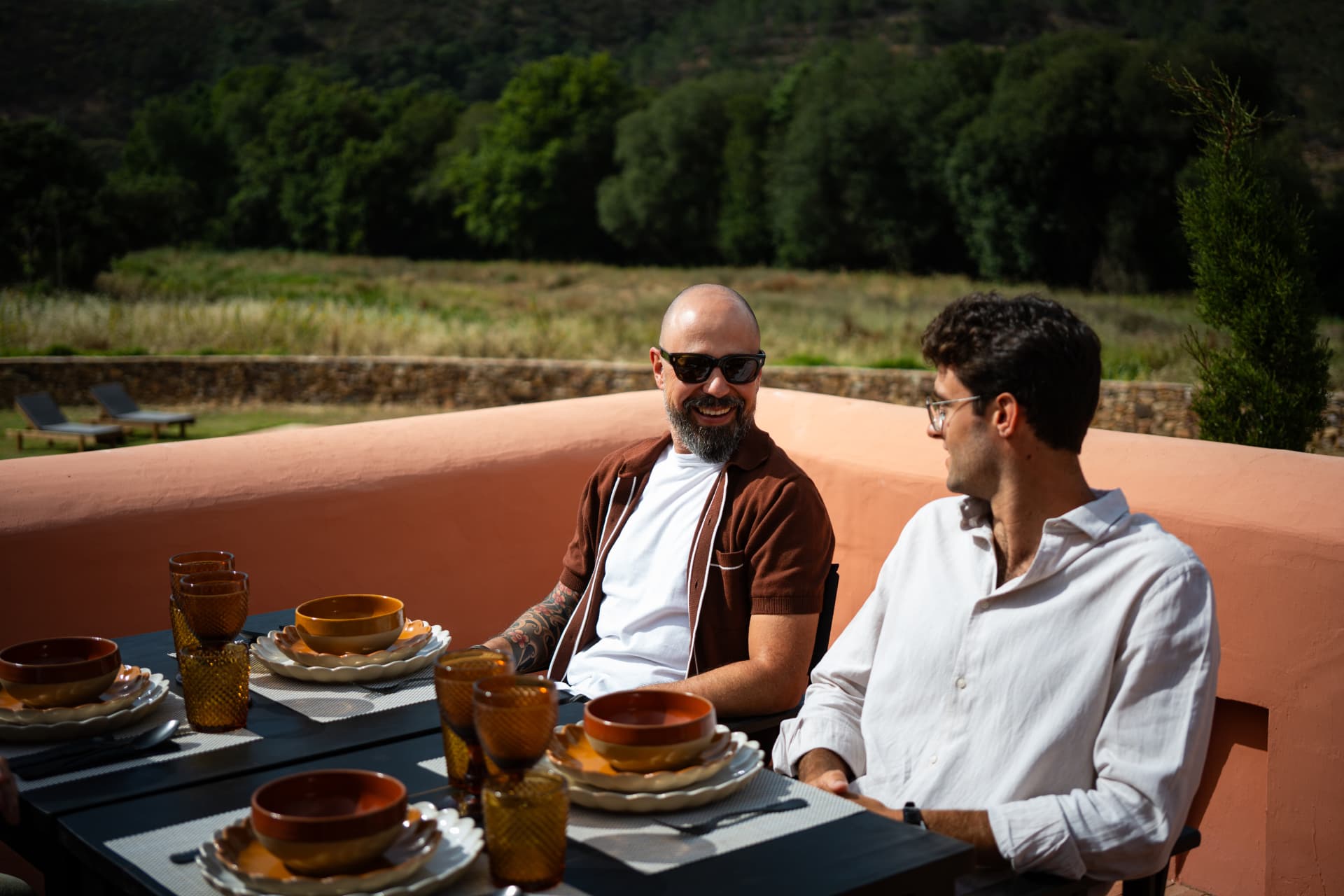 Two guests laughing over lunch at a pink-washed hotel terrace overlooking the valley