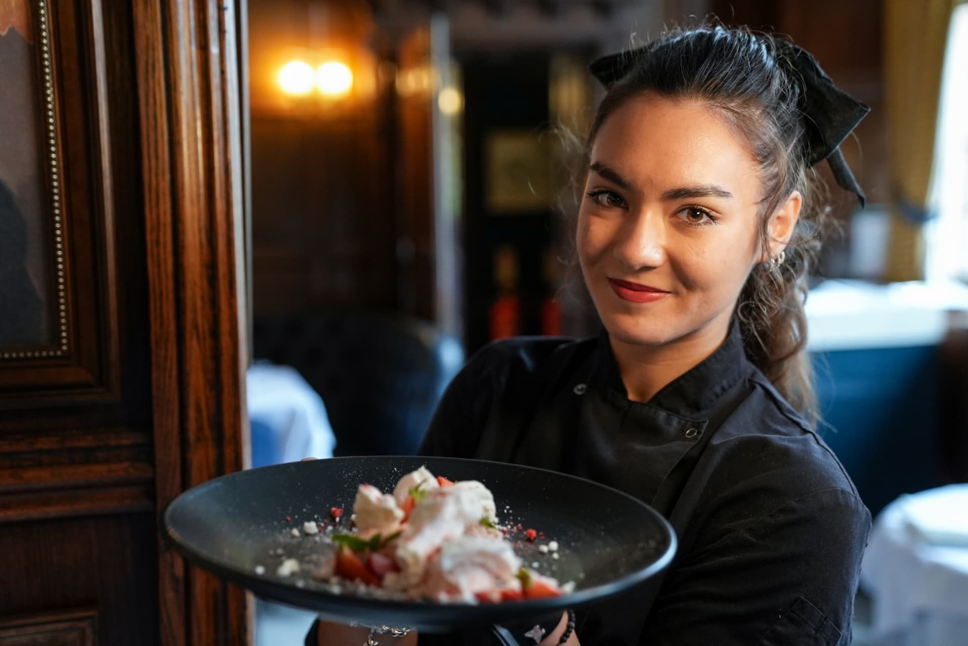 A smiling member of staff presenting a dessert in a wood-panelled dining room