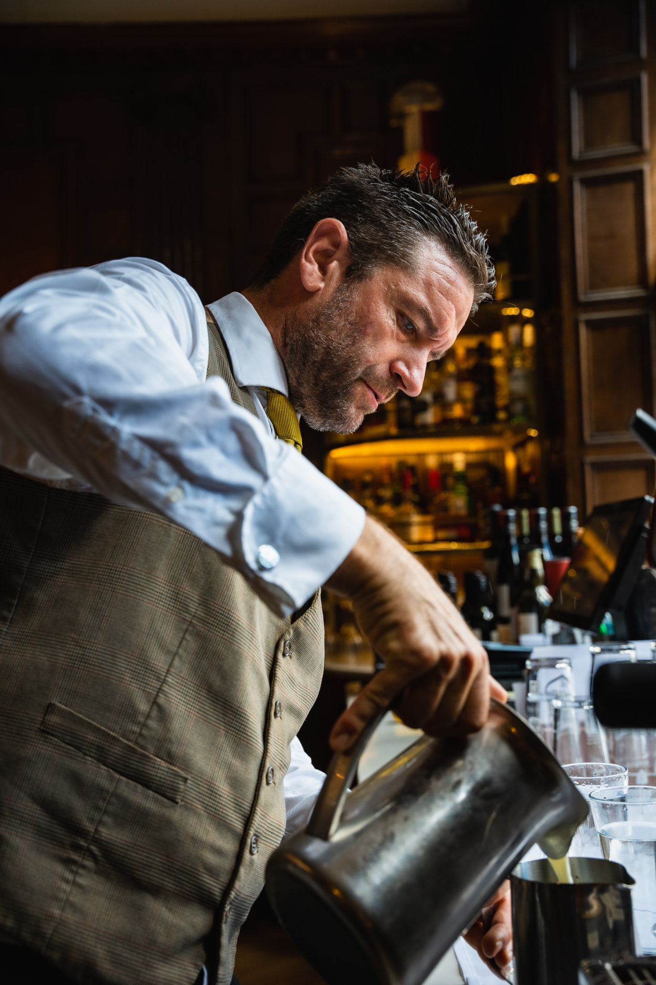 A bartender in a tweed waistcoat carefully pouring a drink at a wood-panelled hotel bar